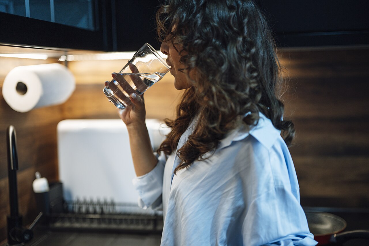 Una mujer de cabello rizado bebe agua de un vaso en una cocina con luz cálida