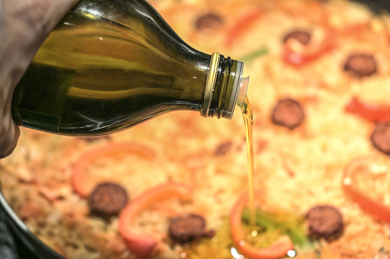 Botella de aceite de oliva sin etiqueta visible vertiendo un hilo de aceite sobre un plato en preparación con tiras de pimiento y rodajas de embutido