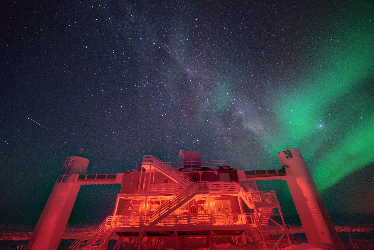 El edificio del laboratorio de IceCube en el Polo Sur, iluminado de rojo bajo un cielo estrellado con la Vía Láctea y una aurora verde