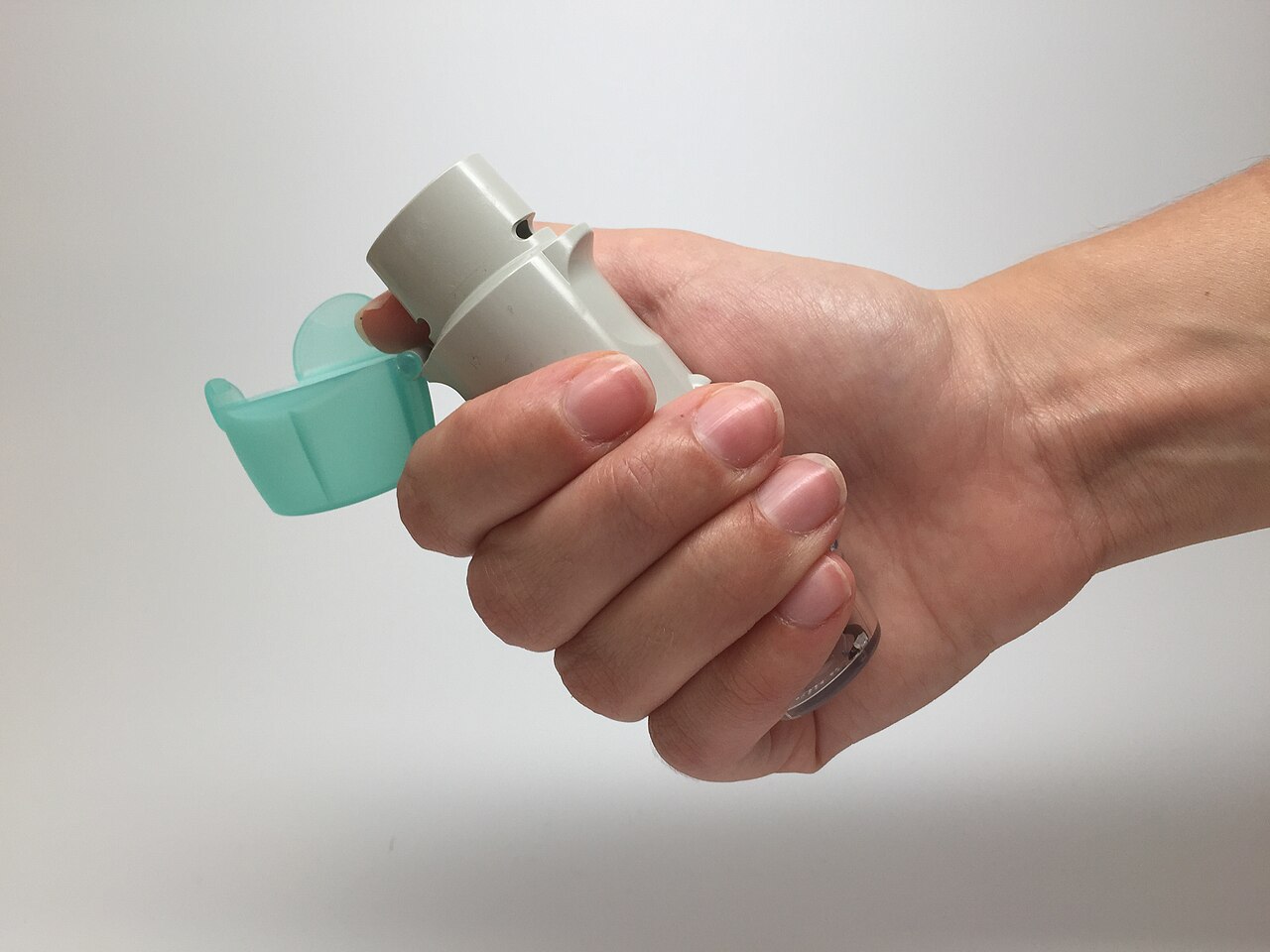 Mano que sostiene un inhalador gris con la tapa verde azulada abierta, sobre un fondo claro