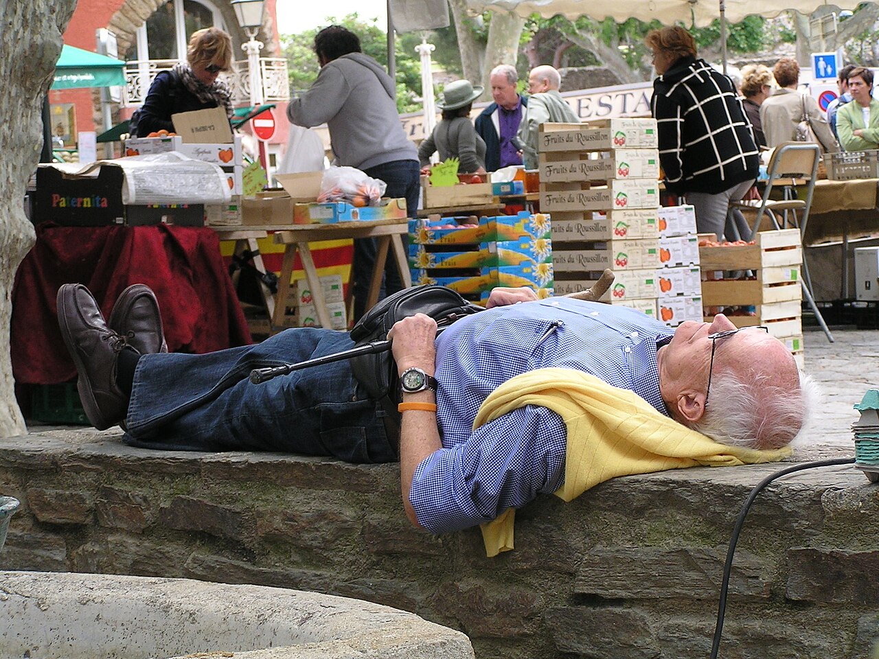 Un hombre mayor duerme la siesta tumbado sobre un muro de piedra junto a los puestos de un mercado