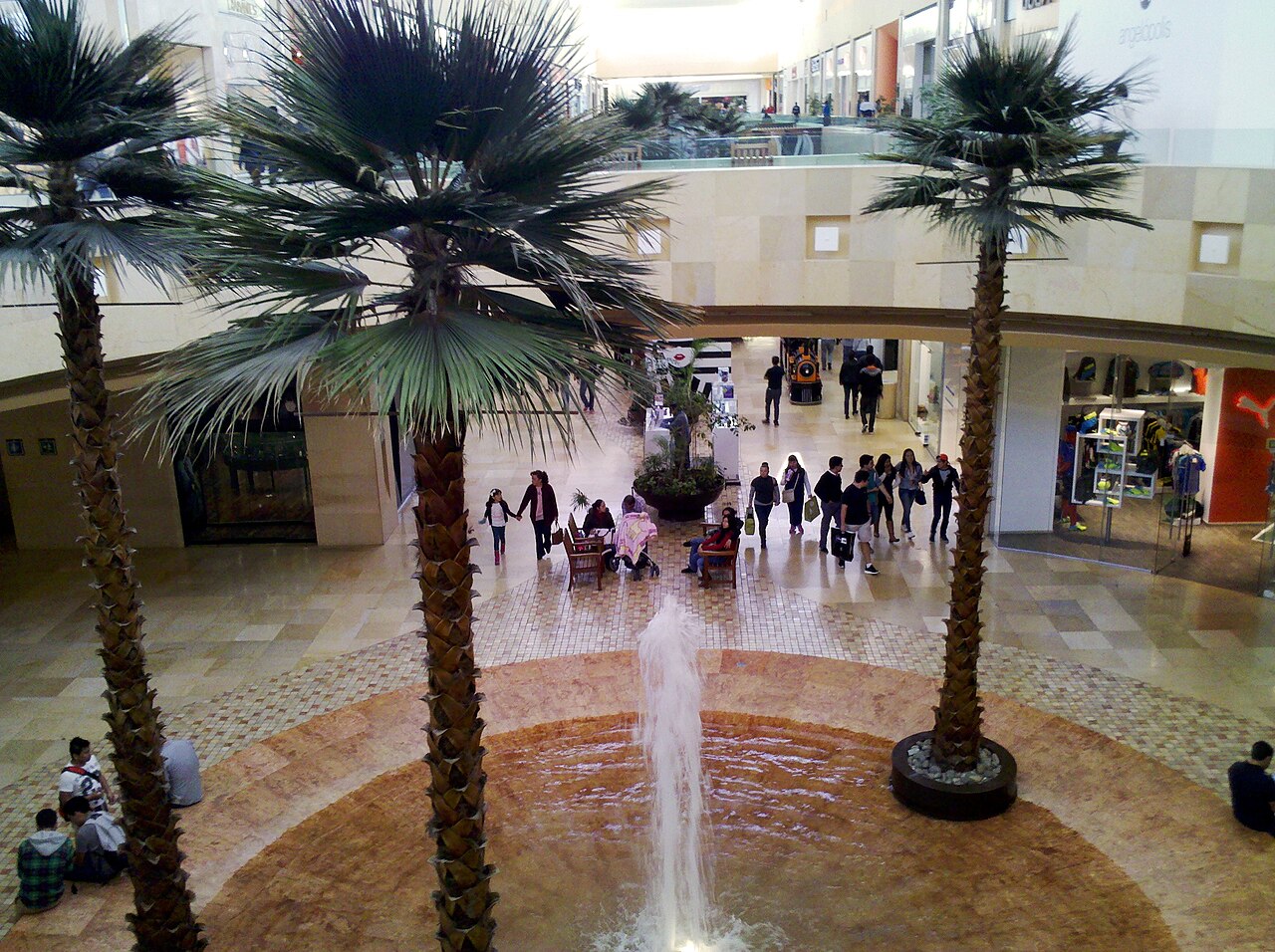 Interior del Centro Comercial Angelópolis, en Puebla, con visitantes caminando entre palmeras y una fuente