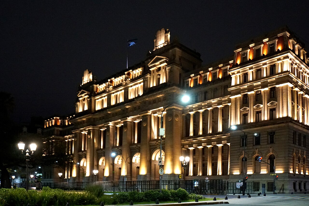 Fachada iluminada de noche del Palacio de Justicia de Buenos Aires, sede de la Corte Suprema de Argentina, con columnas clásicas y una bandera argentina en lo alto