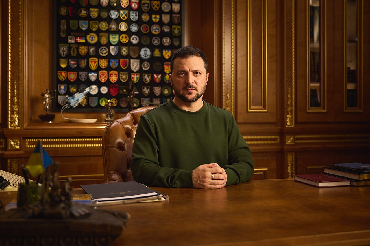 Volodímir Zelenski sentado ante un escritorio de madera, con un panel de parches militares al fondo (imagen de archivo de diciembre de 2023)