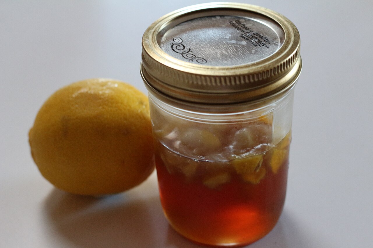 Un tarro de cristal con miel y trozos de limón, junto a un limón entero