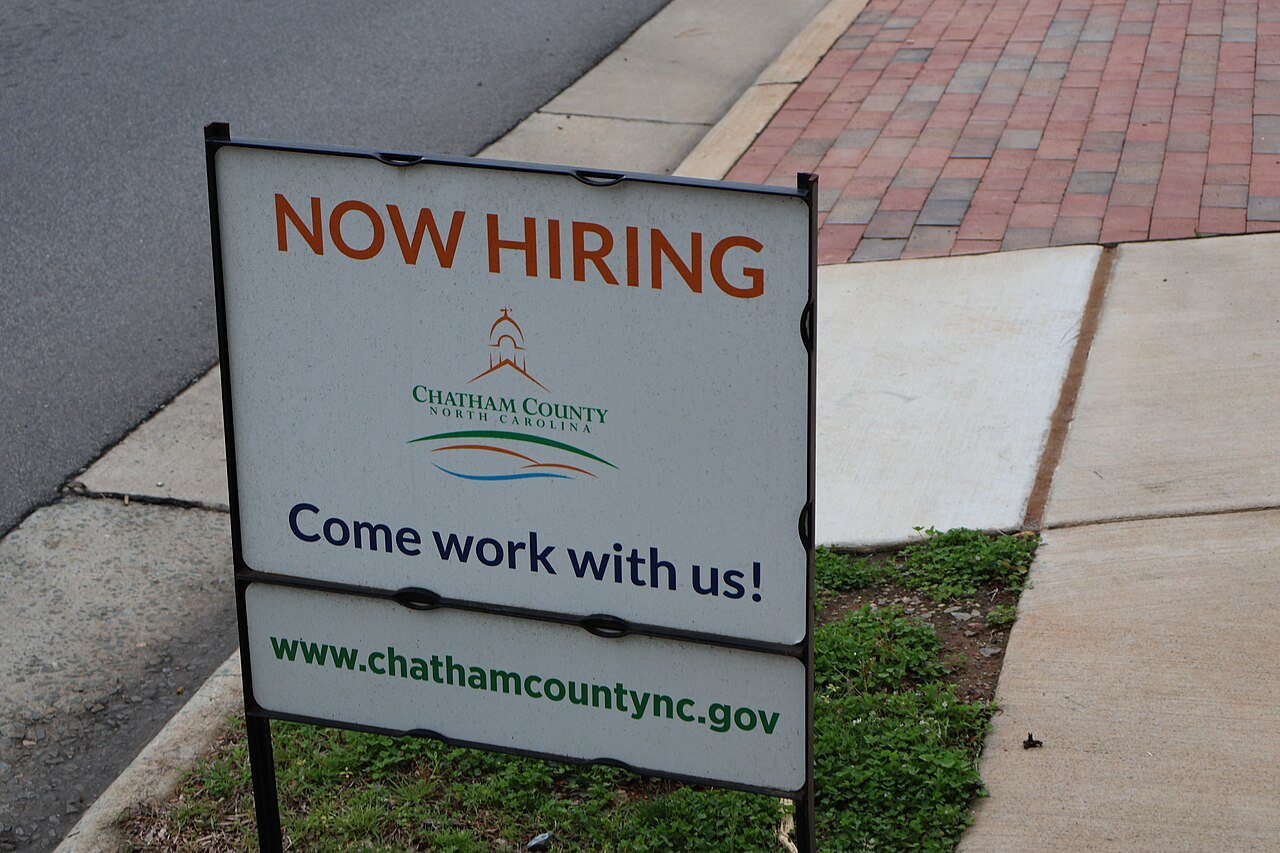 Cartel con el texto “Now hiring” (se busca personal) y la leyenda “Come work with us!” junto a una acera en un condado de Carolina del Norte, en Estados Unidos