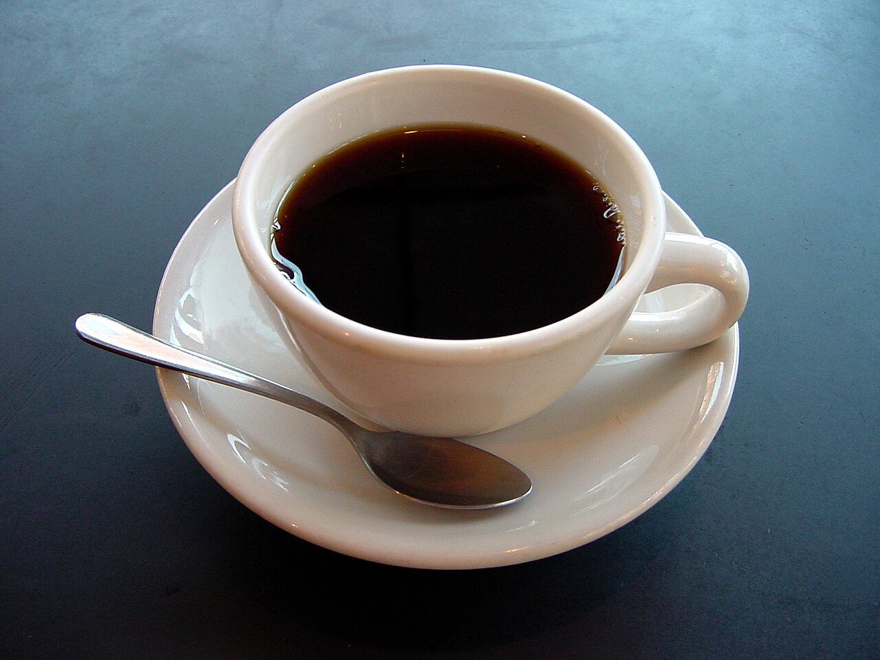 Una taza blanca de café solo sobre un plato, con una cucharita al lado, sobre una mesa oscura