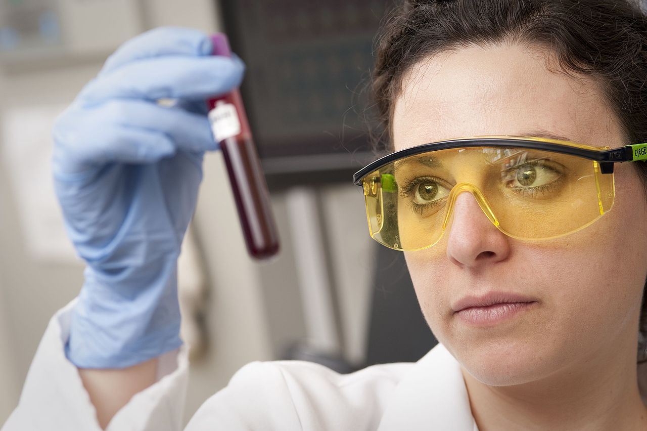 Una técnica de laboratorio con guantes y gafas de protección observa un tubo con una muestra de sangre