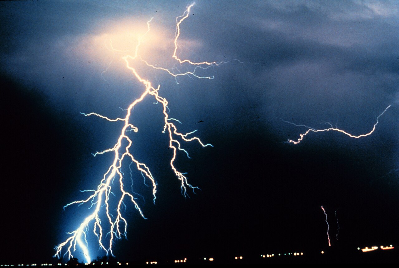 Un rayo con muchas ramificaciones cae desde una nube de tormenta sobre una zona con luces en el horizonte