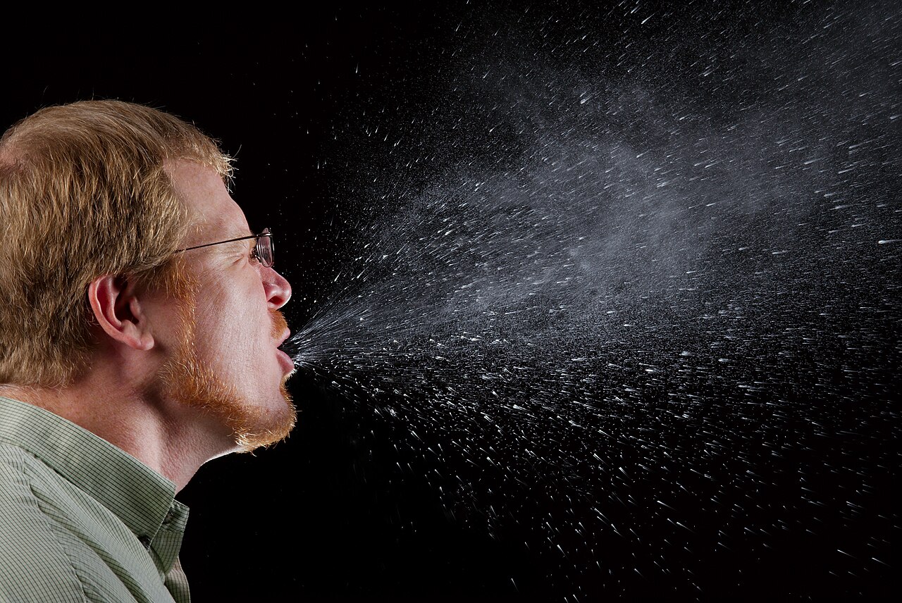 Hombre con gafas estornudando sobre fondo oscuro, con las gotas de saliva visibles en el aire
