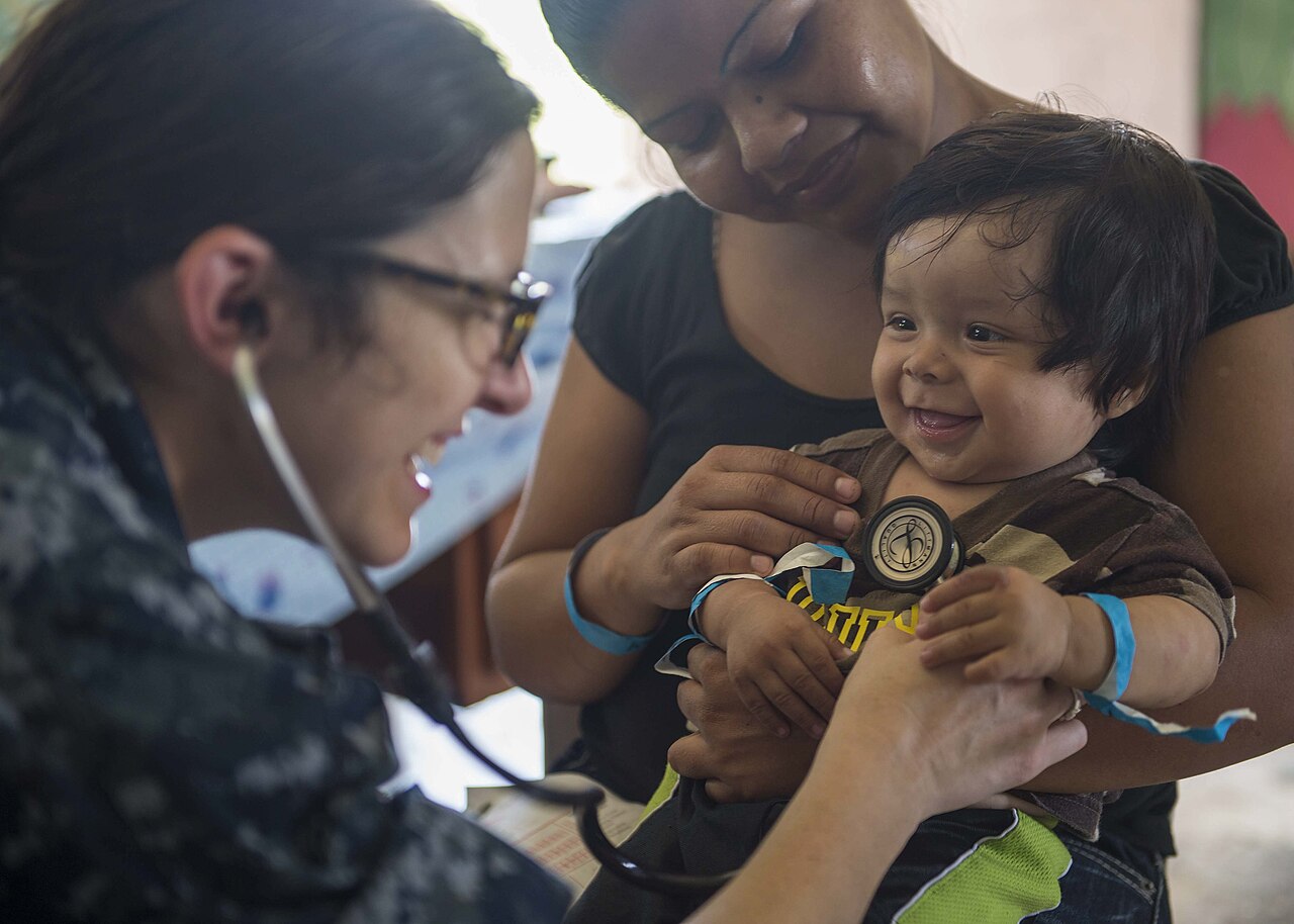 Una pediatra con estetoscopio examina a un bebé sonriente que sostiene su madre