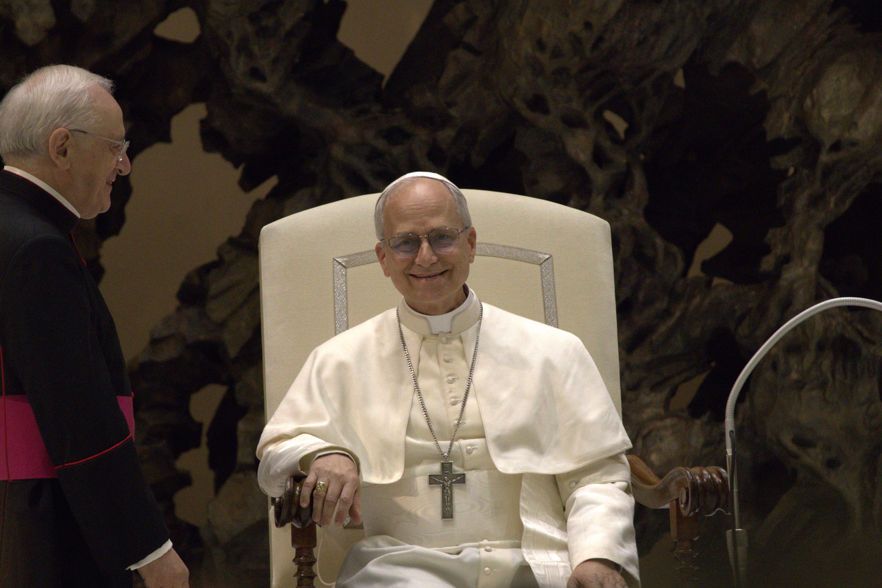 El papa León XIV sonríe sentado en una silla blanca, junto a un clérigo de pie, durante una audiencia con periodistas el 12 de mayo de 2025